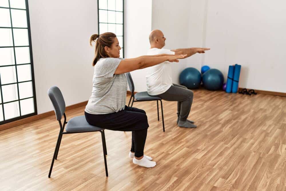 Middle age hispanic couple stretching using chair at sport center. Gym sur chaise - Don Du Souffle
